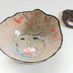 May include: A ceramic bowl with a crackled glaze and a painted face. The bowl is light brown with red, yellow, blue, and white accents.