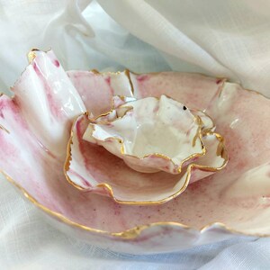 May include: Two nested ceramic bowls with organic, wavy edges and a glossy finish. The bowls feature a white base with soft pink marbled patterns and speckles, accented by thin, metallic gold trim along the irregular, petal-like rims.