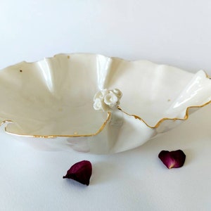 May include: A white porcelain bowl with a ruffled edge and gold trim. A cluster of small white flowers adorns the side. Scattered gold flecks and maroon petals add detail to this decorative piece.