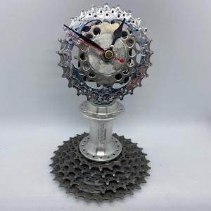 May include: A distinctive clock made from recycled bicycle components. The clock face is a silver gear, with black and red hands. The base is a cluster of gears and a hub, creating a unique industrial design.