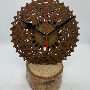 May include: A unique clock crafted from a recycled bicycle gear, with black hands and a red second hand. The clock is mounted on a natural wood base, displaying an industrial design. The clock face has a rusty brown colour, adding to its charm.