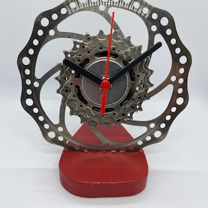 May include: A distinctive clock crafted from recycled bicycle components. It features a silver gear and brake rotor, with black clock hands and a red second hand. The clock is mounted on a red wooden base, adding a vibrant contrast to the industrial design.