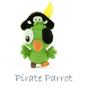 Pode incluir: Um papagaio de crochê verde usando um chapéu de pirata preto com um design de caveira e ossos cruzados e uma pena amarela. O papagaio tem um olho branco e um tapa-olho preto.