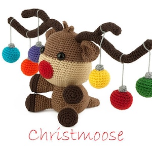 May include: A brown crocheted reindeer with a red nose and antlers holding colorful crocheted ornaments. The reindeer is sitting on a white background. The text "Christmoose" is written in red below the reindeer.