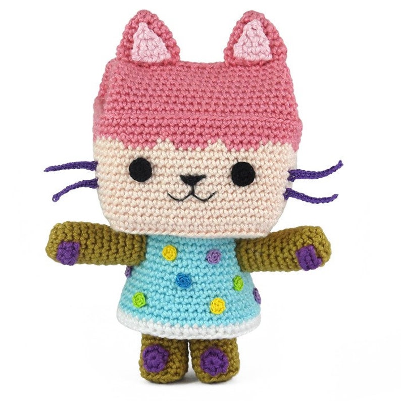 Op de afbeelding: Een gehaakt katten speelgoed met roze oren, een lichtroze hoofd, een lichtbruin lichaam en een blauw-wit geblokte jurk. De kat heeft paarse snorharen en bruine armen en benen.
