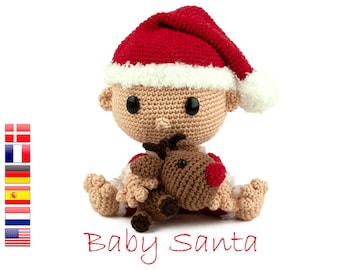 May include: A crocheted baby Santa Claus doll wearing a red and white hat and holding a brown crocheted reindeer. The doll is sitting on a white background.
