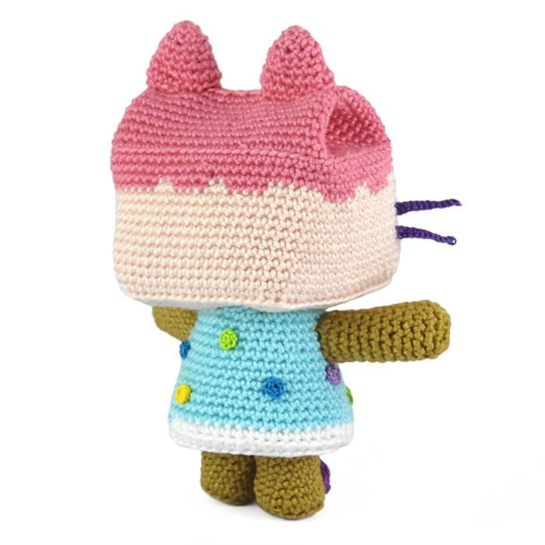 Op de afbeelding: Een gehaakt knuffeldier kat met een roze en witte hoed en een blauwe jurk met kleurrijke stippen. De kat heeft bruine armen en benen.