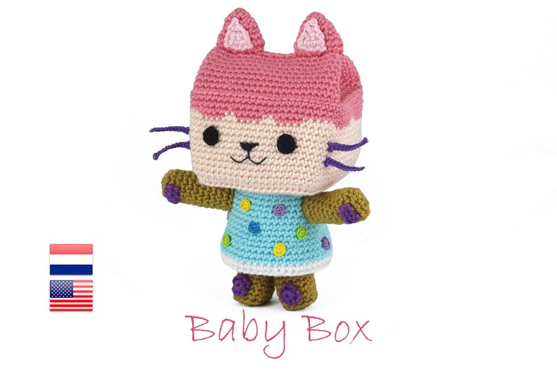 Op de afbeelding: Een gehaakt pluche speelgoed in de vorm van een kat met roze oren, een lichtbruin lichaam en een blauw-wit geblokte jurk. Het speelgoed heeft paarse snorharen en bruine voeten. De tekst "Baby Box" staat onder het speelgoed.