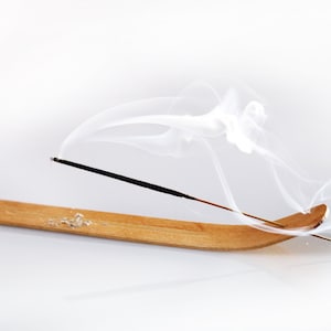 Op de afbeelding: Een houten wierookhouder met een zwarte wierookstok die brandt en witte rook afgeeft. De rook krult en draait in de lucht.