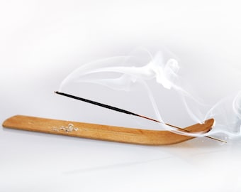 May include: A wooden incense holder with a black incense stick burning and releasing white smoke. The smoke curls and swirls in the air.