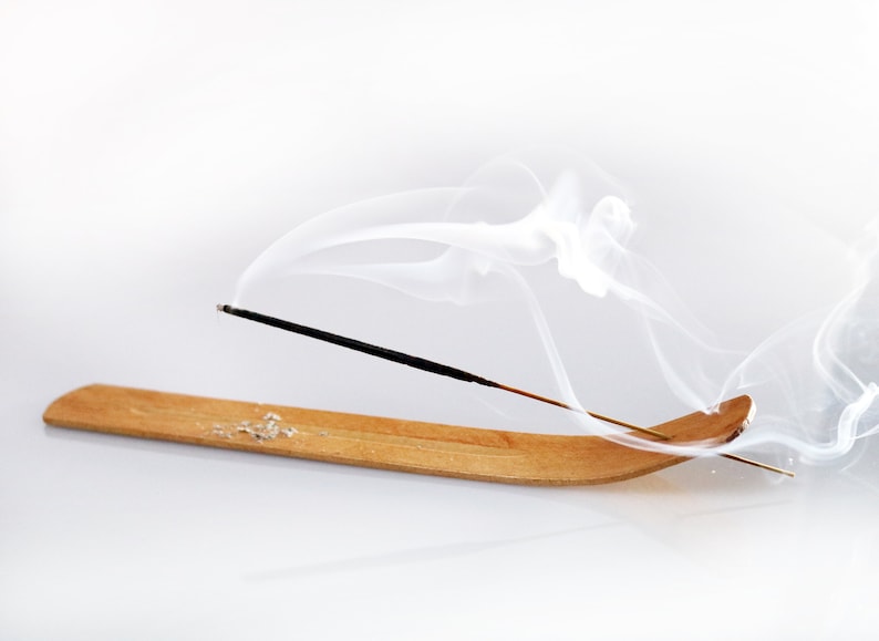 Op de afbeelding: Een houten wierookhouder met een zwarte wierookstok die brandt en witte rook afgeeft. De rook krult en draait in de lucht.