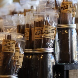 Op de afbeelding: Een assortiment wierookstokjes in doorzichtige plastic zakjes met bruine papieren etiketten. De etiketten bevatten de tekst "Square Scents" en de namen van de geuren, waaronder "Vanille", "Moerbei" en "Wierook & Mirre". De wierookstokjes worden gepresenteerd op een plank met glazen potten.