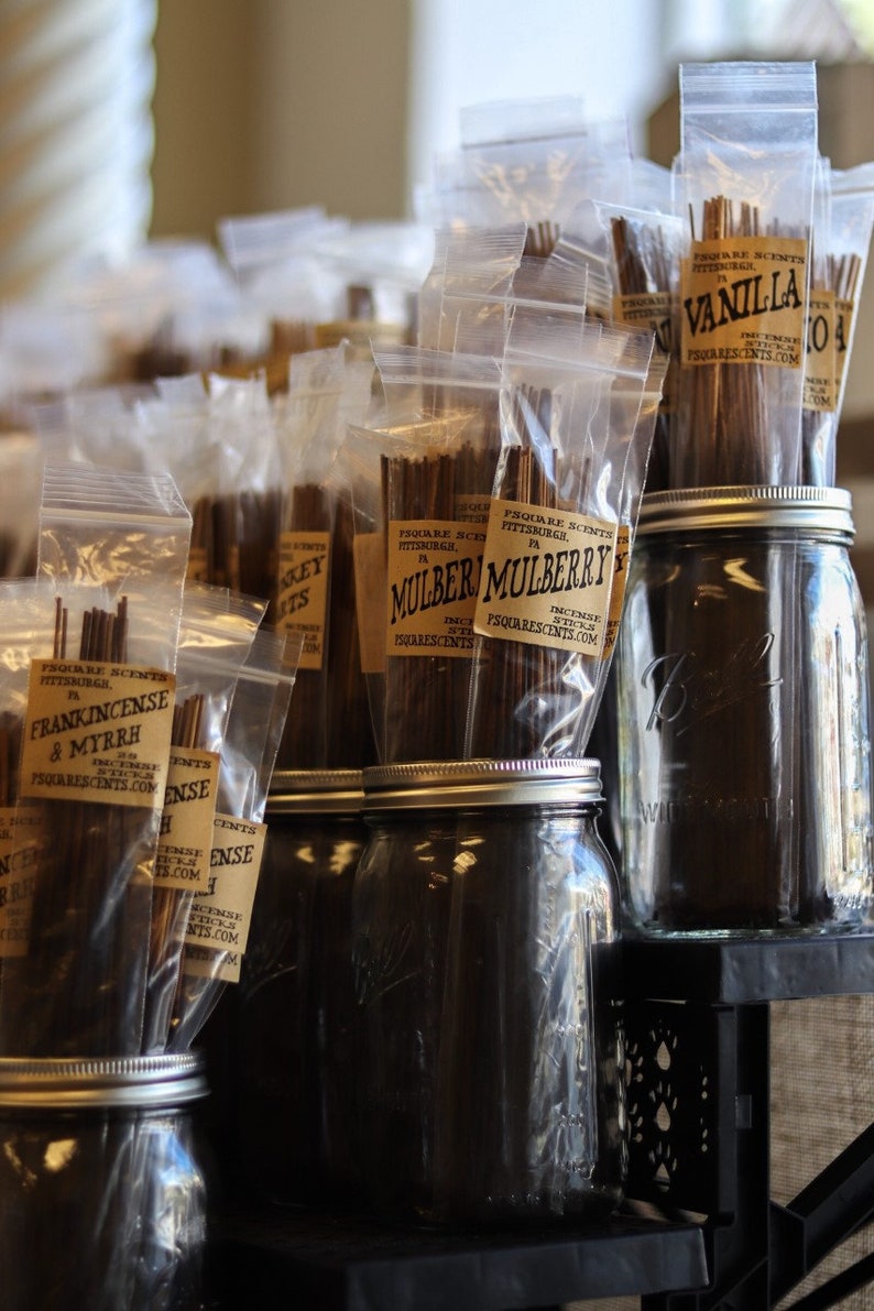 Op de afbeelding: Een assortiment wierookstokjes in doorzichtige plastic zakjes met bruine papieren etiketten. De etiketten bevatten de tekst "Square Scents" en de namen van de geuren, waaronder "Vanille", "Moerbei" en "Wierook & Mirre". De wierookstokjes worden gepresenteerd op een plank met glazen potten.