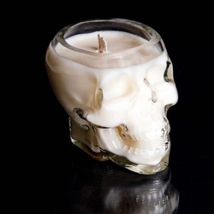 Puede incluir: Un portavelas con forma de calavera hecho de vidrio transparente, lleno de cera blanca. La vela tiene una mecha y está sobre un fondo negro. El diseño de la calavera es detallado, con rasgos faciales visibles.