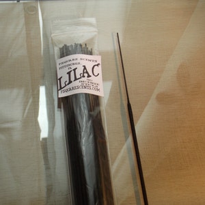 Op de afbeelding: Een pakket zwarte wierookstokjes met een wit label waarop "SQUARE SCENTS PITTSBURGH PA LILAC" en een websiteadres staat. Het pakket ligt op een lichtbruin oppervlak.