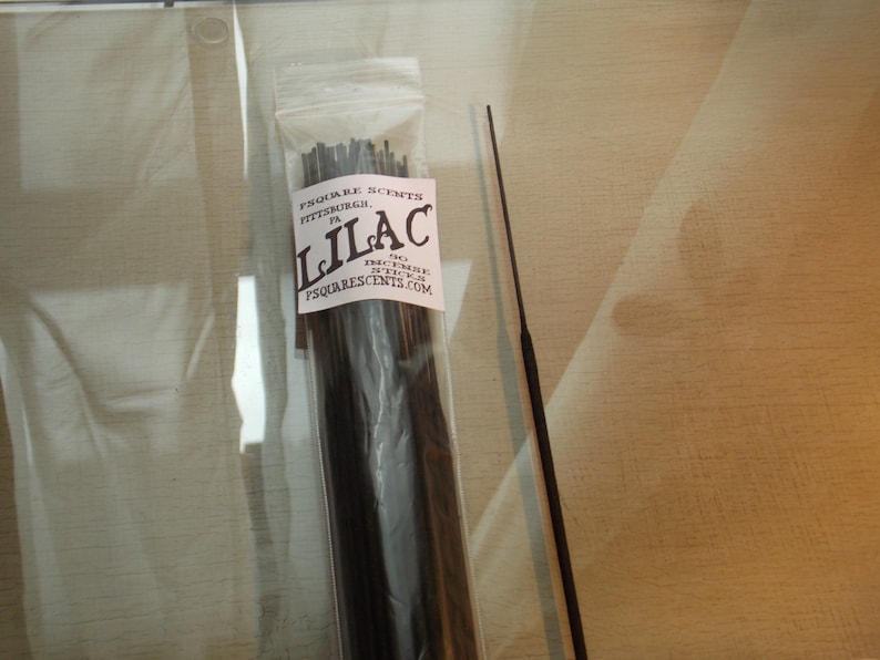 Op de afbeelding: Een pakket zwarte wierookstokjes met een wit label waarop "SQUARE SCENTS PITTSBURGH PA LILAC" en een websiteadres staat. Het pakket ligt op een lichtbruin oppervlak.