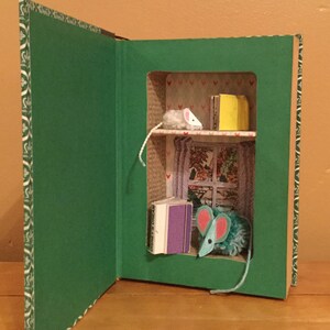 May include: A green book with a small, open door revealing a miniature room with a white mouse, a blue mouse, and books on a shelf. The room has a window with a view of a forest.