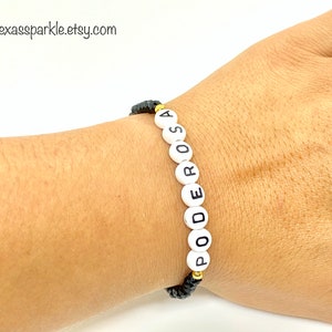 May include: A black string bracelet with white beads that spell out the word "PODEROSA".
