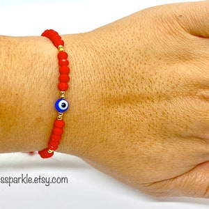 May include: Red beaded bracelet with a blue and white evil eye charm and gold accents. The bracelet is worn on a wrist. The website address alexassparkle.etsy.com is visible at the bottom of the image.