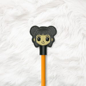 May include: A black and yellow embroidered pencil topper shaped like a cartoon girl's face. The pencil is orange. The topper has a black outline and is stitched with yellow and black thread. The background is white and fluffy.