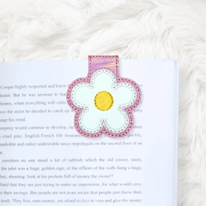 Könnte beinhalten: Ein blütenförmiger Lesezeichen in Weiß und Rosa mit gelbem Zentrum, das auf einem Buch liegt. Das Lesezeichen hat ein gesticktes Design und eine strukturierte Oberfläche. Die Seiten des Buches sind sichtbar, mit gedrucktem Text.