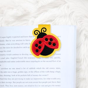 May include: A yellow, red, and black ladybug bookmark. The bookmark is made of felt and is in the shape of a ladybug with black spots on its red wings. The bookmark is inserted into a book.