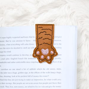 May include: A brown leather bookmark shaped like a cat paw, with a pink paw pad and black stitched details. The bookmark is inserted into a book, with the text of the book visible.