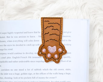 May include: A brown leather bookmark shaped like a cat paw, with a pink paw pad and black stitched details. The bookmark is inserted into a book, with the text of the book visible.