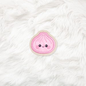 Op de afbeelding: Een roze geborduurde patch in de vorm van een knoedel met een lachend gezicht. Het ontwerp heeft zwarte geborduurde ogen, een kleine gebogen mond en twee roze ronde wangen. De patch heeft een lichtbeige rand met zichtbare stiksels.