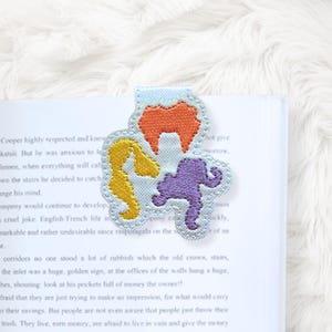 May include: A light blue bookmark with an embroidered design of three cartoon witch hairstyles in orange, yellow, and purple. The bookmark is placed in a book.