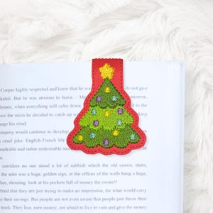 Puede incluir: Un marcapáginas rojo con forma de árbol de Navidad con un diseño de árbol bordado en verde, adornado con adornos coloridos y una estrella amarilla. El marcapáginas está insertado en un libro con texto impreso.