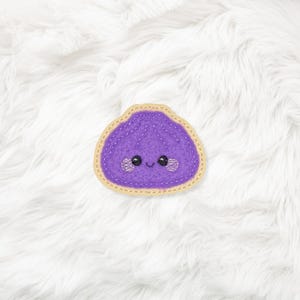 May include: A purple felt dumpling with a smiling face and embroidered details. The dumpling has a light tan border with yellow stitching. The image is set against a white, fluffy background.