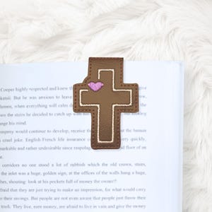 May include: A brown leather cross-shaped bookmark with a pink heart and white stitching. The bookmark is placed on the pages of a book. The bookmark is a religious item.