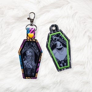 May include: Two coffin-shaped keychains. One features Sally from The Nightmare Before Christmas, with a yellow heart accent. The other keychain has an image of Oogie Boogie. Both keychains have a silver clasp.