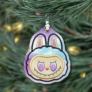 May include: A colorful Christmas ornament with a cartoon monster face design. The ornament features a purple, blue, and yellow color scheme with a smiling face and bunny ears. It hangs from a white loop against a blurred green background.
