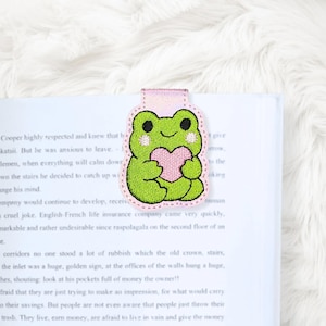May include: A green frog bookmark with a pink heart, embroidered with black details and a pink border. The bookmark is attached to a book with printed text. The bookmark is a cute accessory for book lovers.
