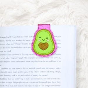 May include: A pink and green avocado-shaped bookmark with a smiling face and a brown heart. The bookmark is placed on a book with printed text. The bookmark is made of fabric and has a stitched design.