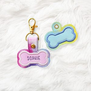 Könnte beinhalten: Ein schlüsselanhänger und ein Haustieranhänger in Knochenform. Der Schlüsselanhänger hat einen rosa, lila und blauen Knochen mit dem Namen "SOPHIE" aufgestickt. Der Anhänger ist hellblau mit gelben und blauen Verzierungen. Beide liegen auf einer weißen, flauschigen Oberfläche.