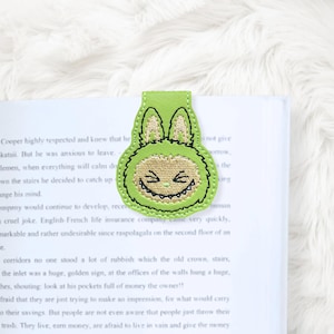 May include: A green and tan embroidered bookmark in the shape of a cartoon monster with a smiling face and bunny ears. The bookmark is made of a soft material and is placed on the pages of a book.