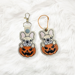 May include: Two embroidered keychains featuring a French bulldog design. The dog is cream-colored and peeking over a carved orange pumpkin. One keychain has a silver clasp, and the other has a gold chain.