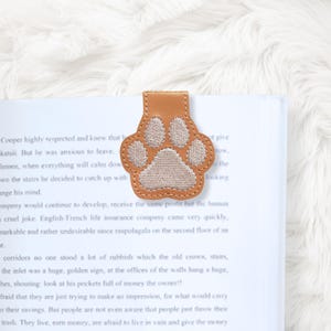 May include: A tan leather bookmark shaped like a paw print, with embroidered details. The bookmark is placed on the page of a book with printed text. The paw print design is a charming accessory for book lovers.