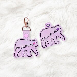 May include: Two lavender bear-shaped keychains with the word "mama" in black script. One keychain has a metal clasp and a pink button. A small pink heart is next to the word "mama" on each bear.