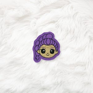May include: A purple felt patch featuring an embroidered cartoon face with a bun and braid hairstyle. The face has large black eyes, a small smile, and a tan skin tone. The patch is on a white, fluffy surface.