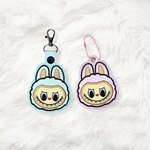 May include: Two embroidered keychains in the shape of a cartoon bunny face. One is light blue with a silver clasp, and the other is pink with a pink chain. Both have a yellow face, brown ears, and a black outline.