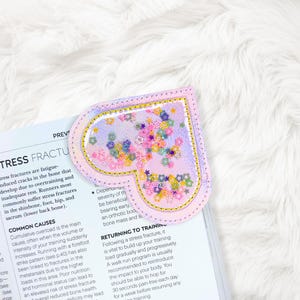 May include: A pink heart-shaped bookmark with a clear, glitter-filled center and floral embellishments. The bookmark is placed on top of an open book. The bookmark has a gold trim.