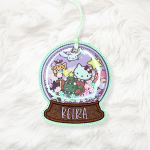 May include: A snow globe-shaped Christmas ornament featuring Hello Kitty and other characters. The ornament has a clear front, a purple background, and a green border. The name "KEIRA" is displayed on the base.