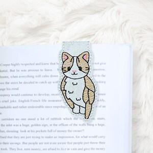 May include: A cat-shaped bookmark with a white, tan, and pink embroidered design. The bookmark is placed on the pages of a book. The cat has a smiling face and is outlined in black. The bookmark is a cute accessory for book lovers.