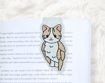 May include: A cat-shaped bookmark with a white, tan, and pink embroidered design. The bookmark is placed on the pages of a book. The cat has a smiling face and is outlined in black. The bookmark is a cute accessory for book lovers.