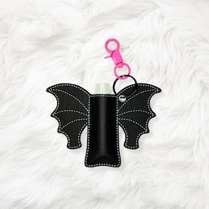 May include: Black bat-shaped lip balm holder with white stitching and a pink clip. The holder has a black ring and a pink clip for attaching to a keychain or bag. The holder is designed to hold a lip balm tube.