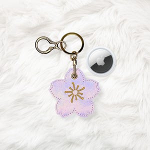 以下が含まれることがあります： 金色のステッチと金色のリングと留め金が付いた、薄紫色の花の形をしたキーホルダー。 Apple AirTagがキーホルダーの隣にあります。 キーホルダーは白いふわふわの表面にあります。